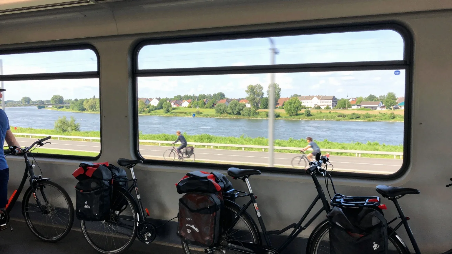 Ein Reiserad mit Packtaschen im Fahrradabteil eines Regionalzugs, draußen zieht eine grüne Flusslandschaft vorbei